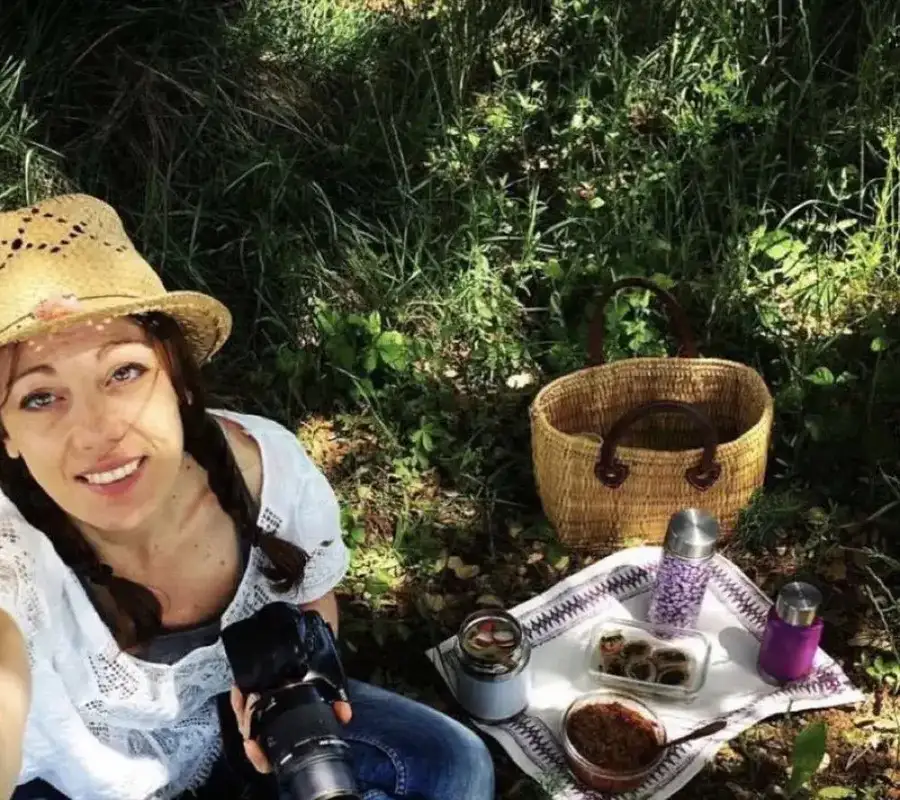 Frédérique Barral assise dans l'herbe, appareil photo à la main, à côté d'un panier et de préparations maison